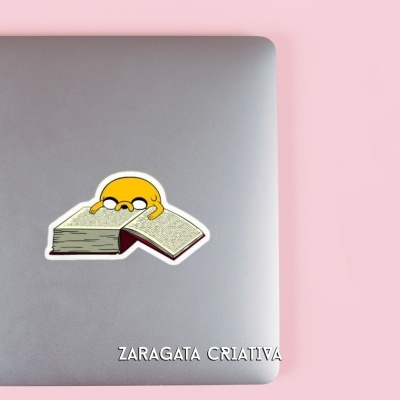 Computador portátil com autocolante de personagem lendo livro em mesa rosa