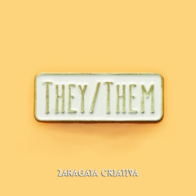 Pin metálico branco com texto THEY/THEM em dourado sobre fundo amarelo.