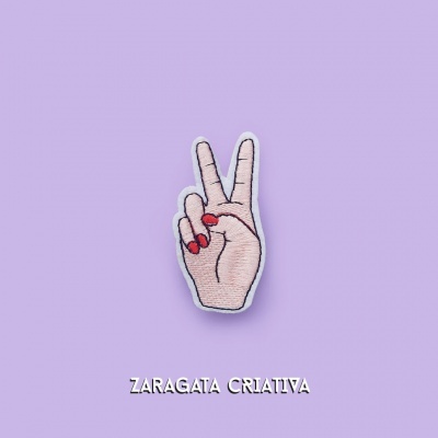 Patch bordado de mão com unhas vermelhas a fazer o gesto de paz sobre fundo lilás com texto ZARAGATA CRIATIVA.
