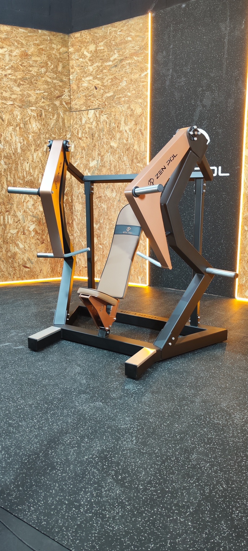 Máquina de treino de peito com acolchoamento bege e estrutura metálica preta e marron numa sala com piso antiderrapante.