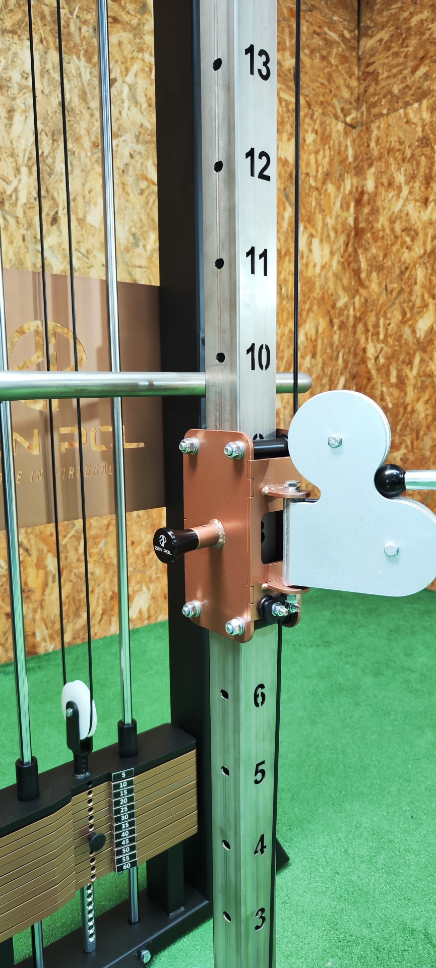 Máquina de exercício com barra ajustável em metal e pesos numa sala com chão verde e parede de OSB.