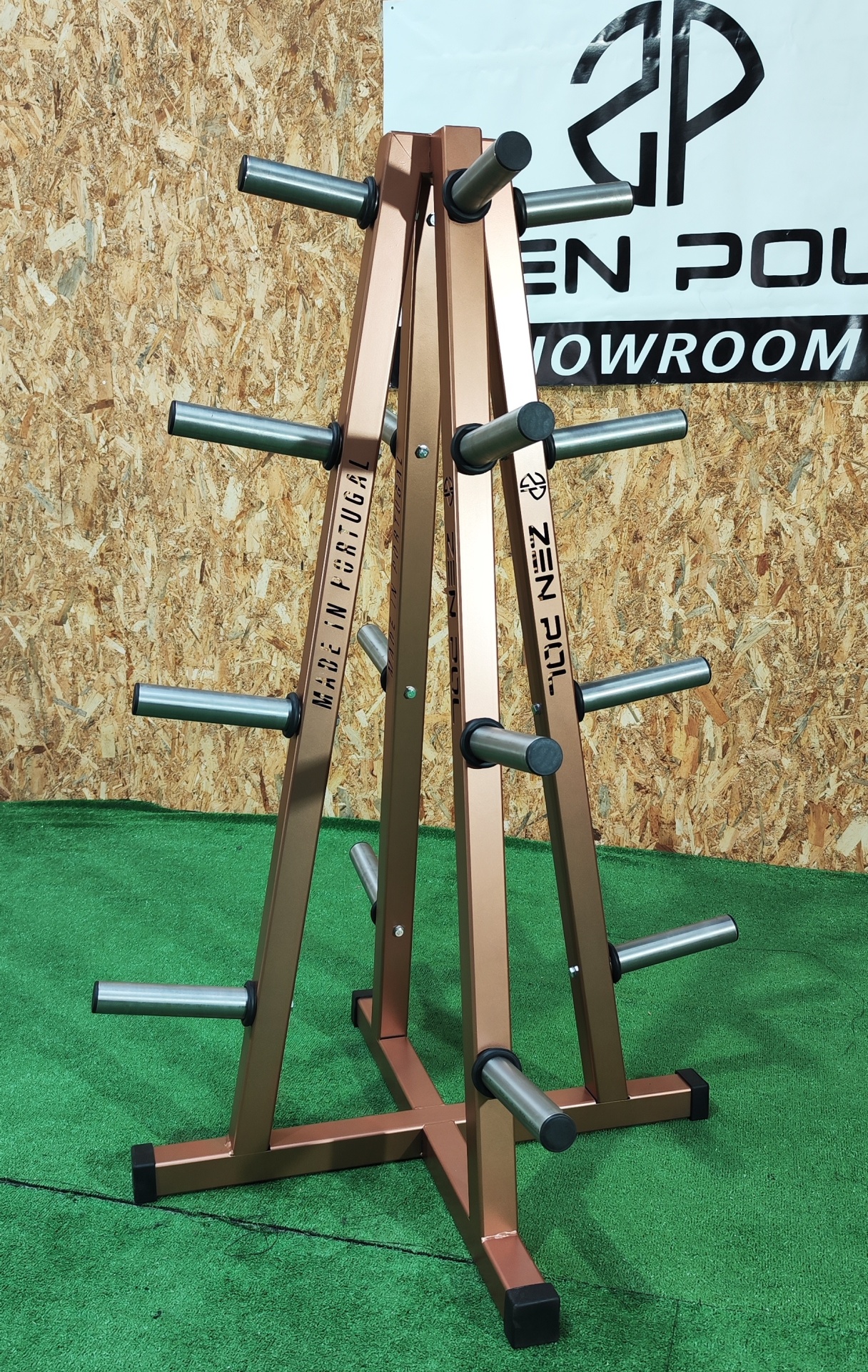 Suporte de pesos bronze com braços azuis num chão verde e fundo de madeira e cartaz