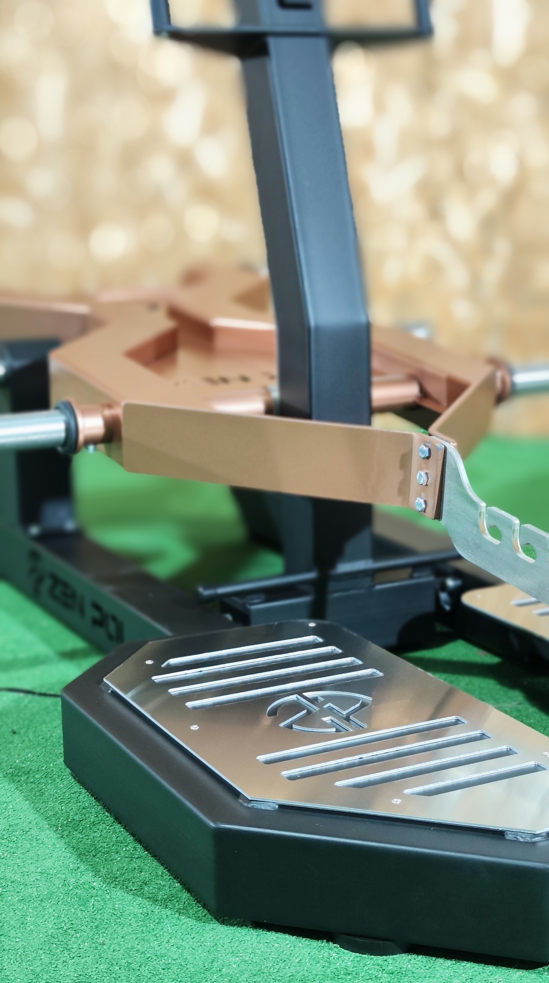 Equipamento de exercício de ginásio com base metálica prateada e estrutura em metal preto e dourado sobre tapete verde