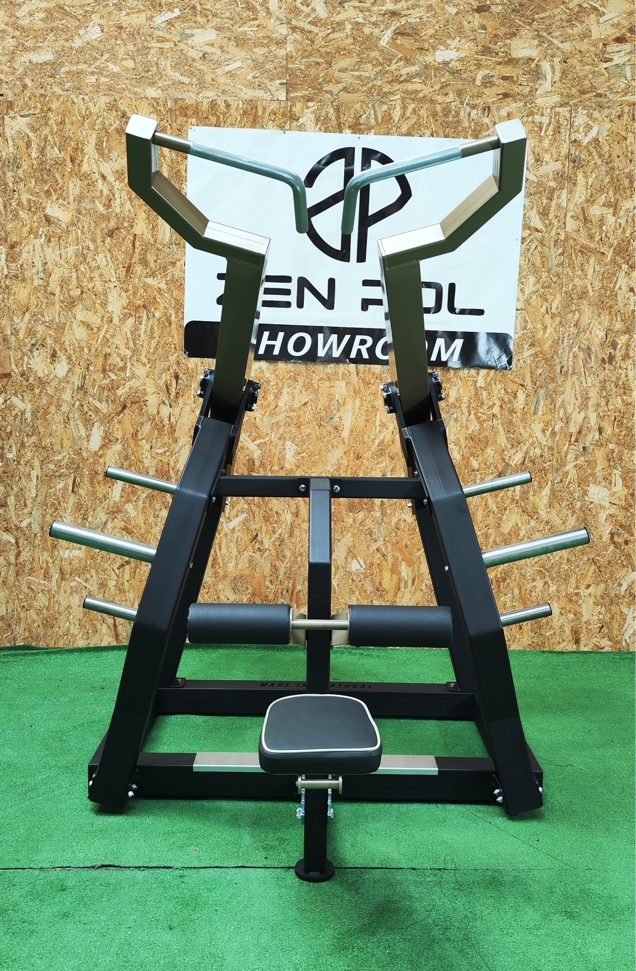 Aparelho de musculação preto com assento cinza em ambiente com tapete verde e parede de madeira
