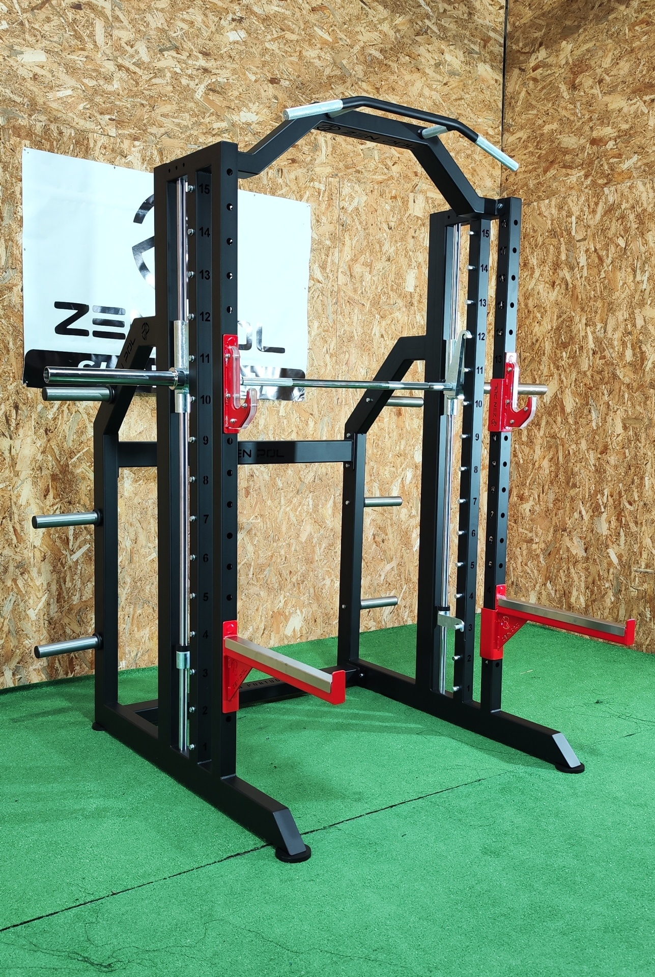 Power rack preto com detalhes vermelhos e barra cromada sobre piso verde e parede de madeira.