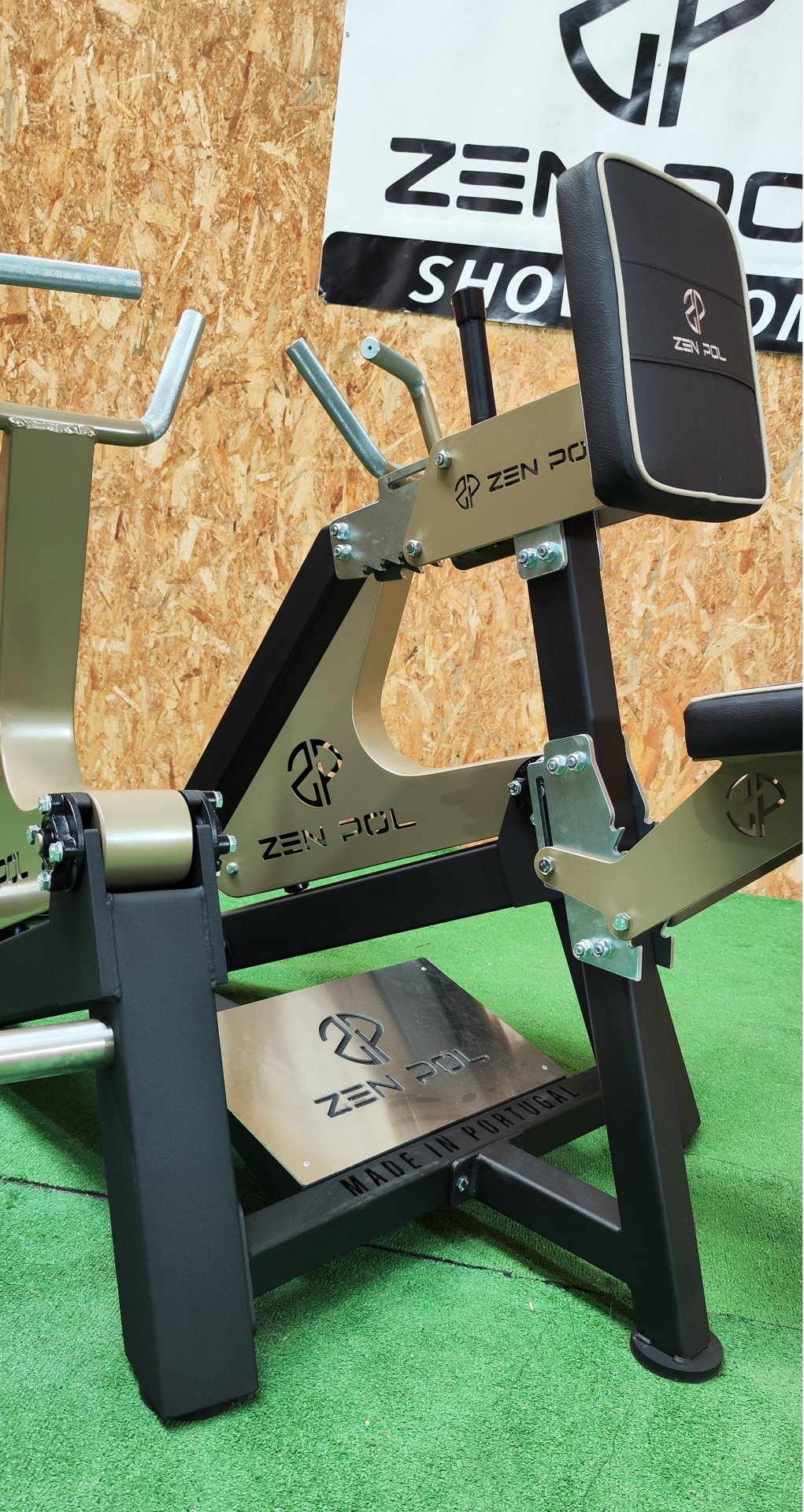 Equipamento de ginásio Zen Pol preto e dourado com assento e encosto, sobre relva artificial e fundo de madeira