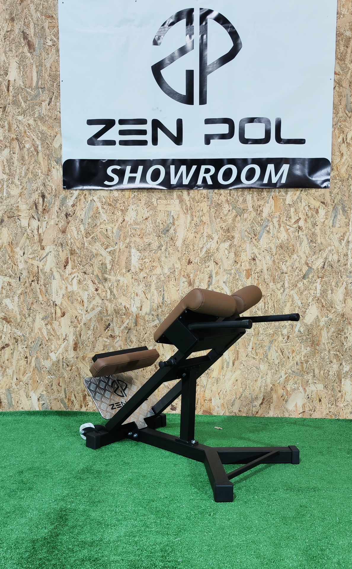 Cadeira de exercício com estrutura metálica preta e elementos acolchoados castanhos, sobre piso verde e parede de madeira aglomerada com cartel 'ZEN POL SHOWROOM'