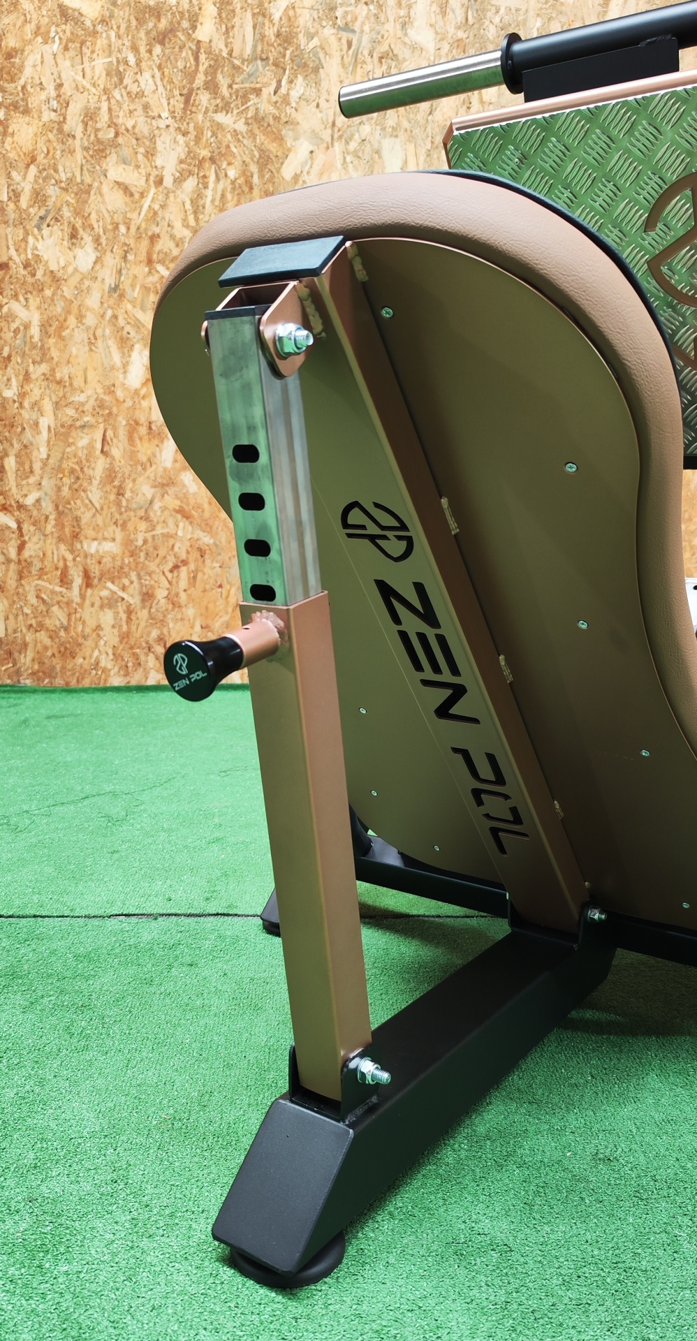 Equipamento de fitness metálico com estofado castanho e texto 'ZEN FIT' numa sala com tapete verde e parede de madeira