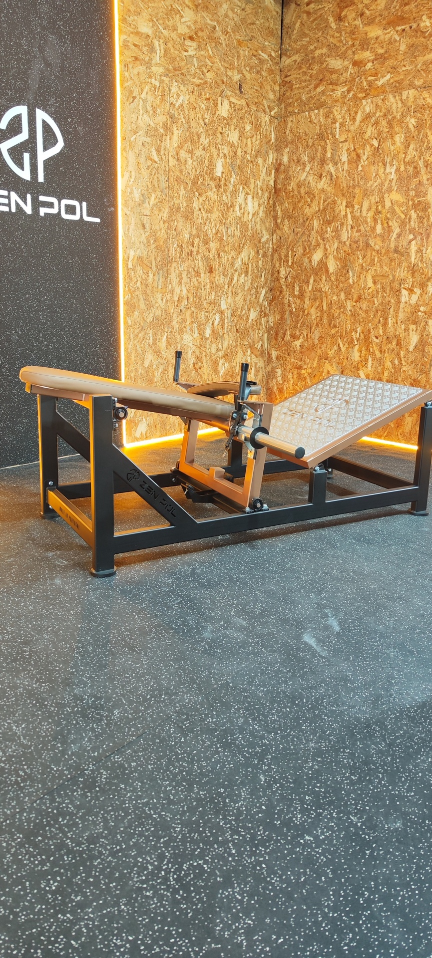 Aparelho de fitness para pernas preto e bege junto a paredes de madeira e parede preta com texto ZEN POL