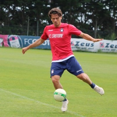 Jogador de futebol com camisola vermelha e calções azuis controla bola num campo de relva