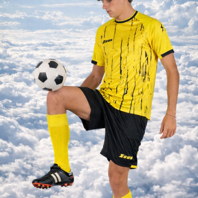 Jogador de futebol com vestuário amarelo e preto controlando bola sobre fundo de céu com nuvens