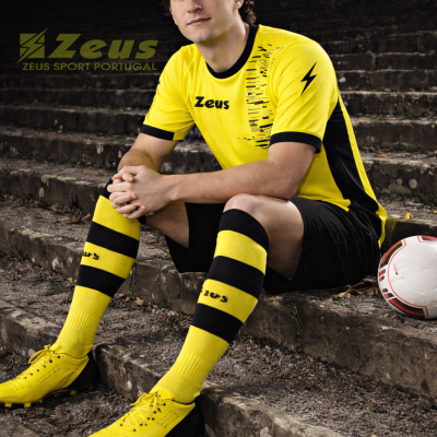 Homem sentado em escadas com equipamento de futebol amarelo e preto da marca Zeus ao lado de uma bola