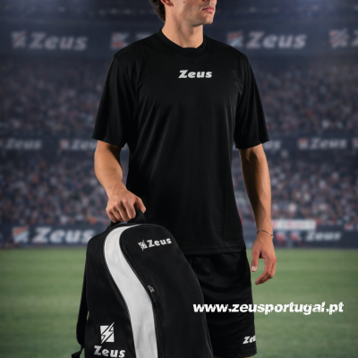 Jovem com roupa desportiva preta e mochila Zeus num estádio de futebol.