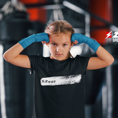 Criança com t-shirt preta 'Zeus' e luvas azuis em ginásio com saco de boxe.