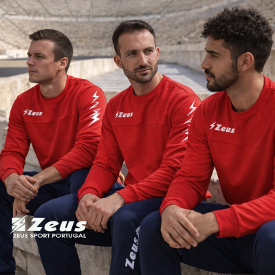 Três homens sentados em estádio usam roupa desportiva vermelha e azul com logo Zeus
