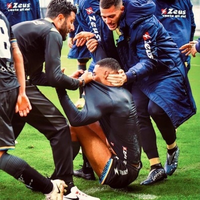 Jogadores e técnicos de futebol em campo com roupa desportiva preta e casacos azuis escuros com texto nas costas