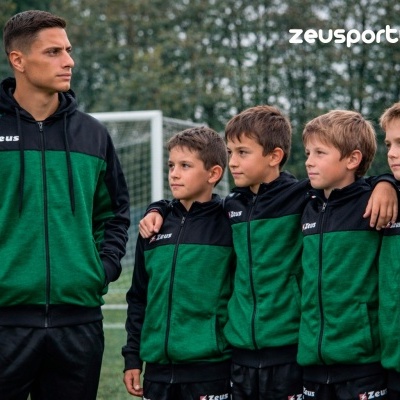 Adulto e quatro crianças vestem conjunto de treino verde e preto com logótipo Zeus, em campo de futebol.