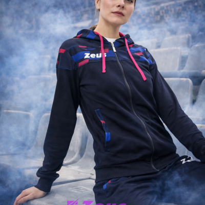 Mulher com casaco desportivo azul escuro com capuz e detalhes coloridos sentada em banco de estádio com fumos azuis ao redor