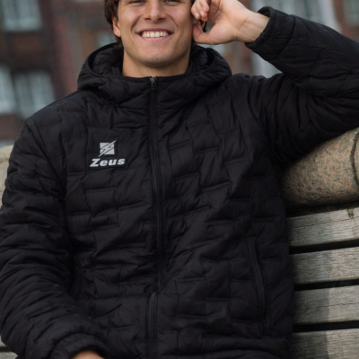 Homem com casaco preto acolchoado com logótipo Zeus sentado num banco de madeira.