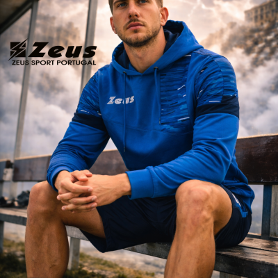 Homem com hoodie azul e calções desportivos azuis sentado em banco com logo Zeus ao fundo