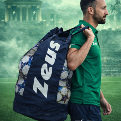 Homem com saco da marca Zeus cheio de bolas de futebol num campo de relva
