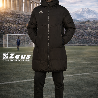 Homem a usar casaco acolchoado preto comprido com capuz e fechos, em campo de futebol com estádio e ruínas antigas ao fundo.