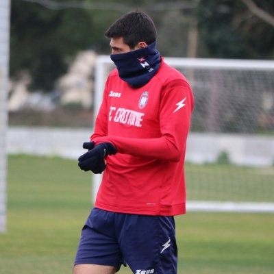 Jogador em campo veste camisola vermelha FC CROTONE, calções e cachecol azuis escuros.