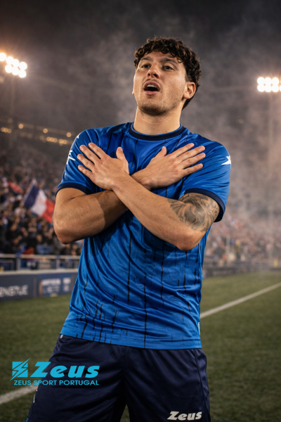 Jogador de futebol com camisola azul e calções azul escuro da marca Zeus em estádio iluminado