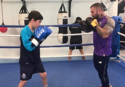 Duas pessoas a praticar boxe num ringue azul com cordas, uma com luvas azuis e outra com luvas amarelas.