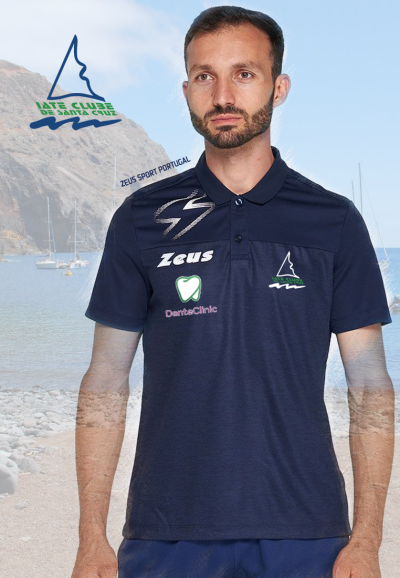 Homem com polo azul escuro com golas e logótipos em ambiente de praia com mar e barcos