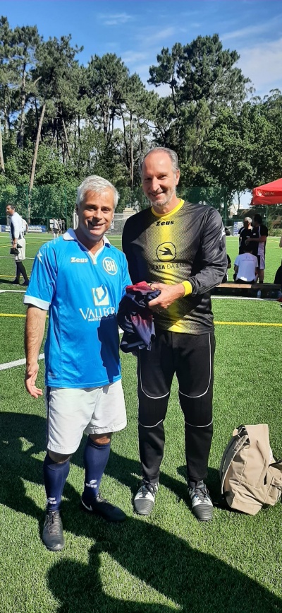 Dois homens em campo de futebol com roupas desportivas, camisolas azul e preta, árvores e tenda vermelha ao fundo