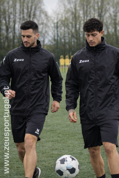Dois homens com fato de treino preto da marca Zeus em campo de futebol com bola preta e branca