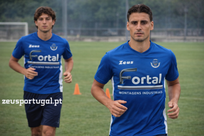 Dois homens a correr com camisolas azuis e calções pretos num campo de futebol.
