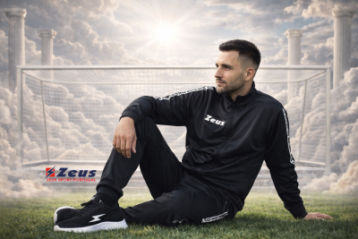 Homem sentado com fato desportivo preto e sapatilhas negras com símbolo branco num campo de futebol com baliza e colunas clássicas ao fundo