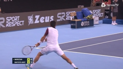 Jogador de ténis em ação numa quadra azul com publicidade e placar visível.