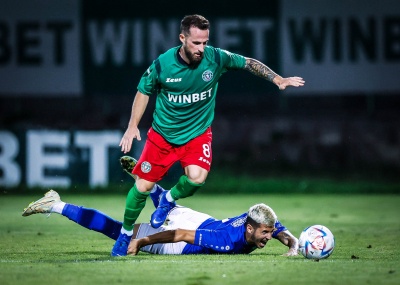 Dois jogadores de futebol em ação num relvado, um de verde e vermelho e outro de azul caído no chão perto da bola.