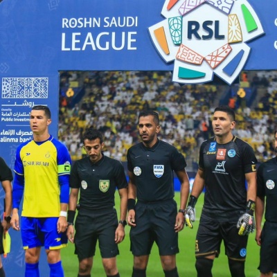 Árbitros e jogadores de futebol em campo com roupa preta e azul-amarela, fundo com texto ROSHN SAUDI LEAGUE