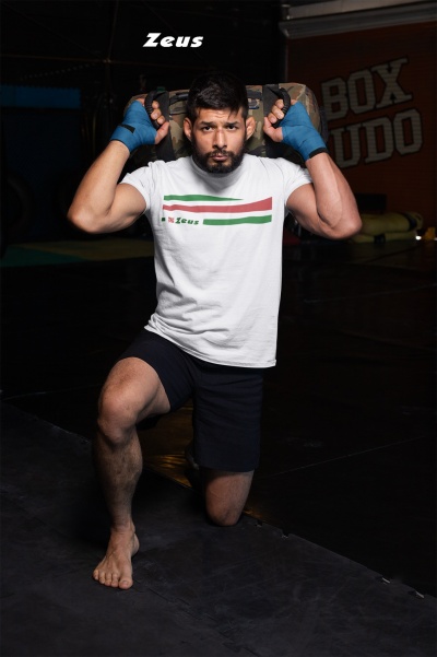 Homem com t-shirt Zeus e calções pretos no ginásio segurando saco de areia