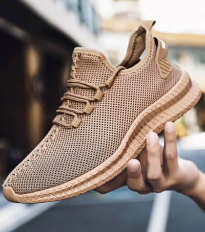 Sapatilha desportiva bege em malha com sola texturada, sendo segurada por uma mão.