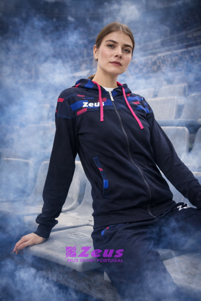 Mulher com casaco desportivo azul escuro com capuz e detalhes coloridos sentada em banco de estádio com fumos azuis ao redor