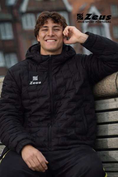 Homem com casaco preto acolchoado com logótipo Zeus sentado num banco de madeira.