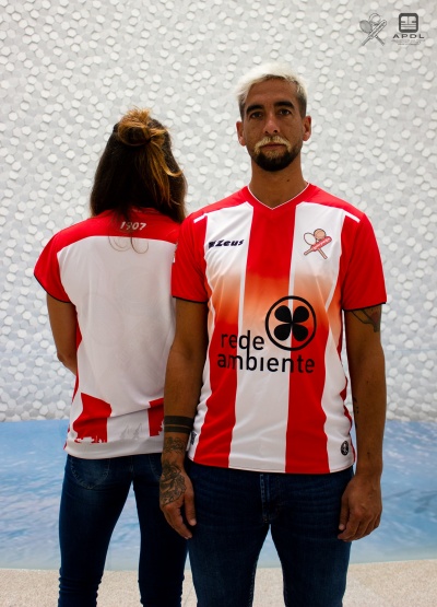 Duas pessoas vestem camisolas desportivas vermelhas e brancas com logótipos e texto, em fundo claro com padrão circular.