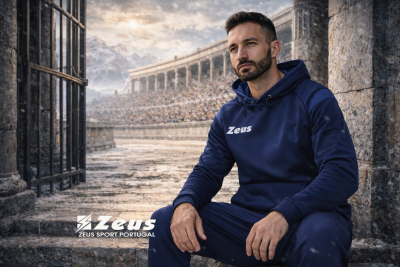 Homem sentado com roupa desportiva azul escura com capuz e logótipo Zeus em arena de pedra antiga