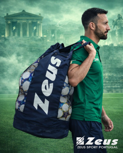 Homem com saco da marca Zeus cheio de bolas de futebol num campo de relva