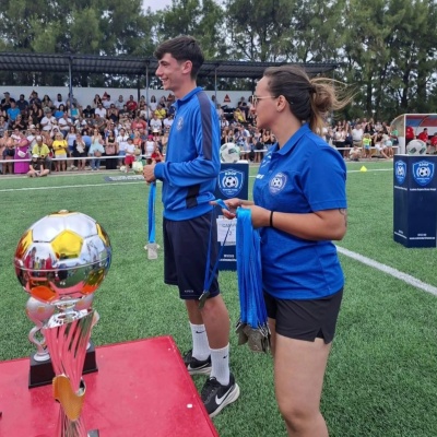Monitores de futebol a distribuir medalhas num campo de relva, com troféu em destaque e plateia ao fundo